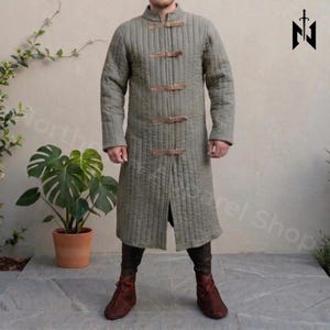 May include: An olive-green quilted coat with brown leather straps. The coat features a high collar and is worn with brown boots. This long coat is inspired by medieval designs, suitable for historical reenactment or costume events.