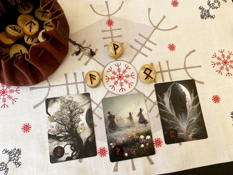 Puede incluir: Un pa&ntilde;o blanco con un patr&oacute;n rojo y gris. Tres cartas con im&aacute;genes de un &aacute;rbol, un grupo de personas bailando y una pluma. Cuatro runas de madera con s&iacute;mbolos est&aacute;n colocadas en el pa&ntilde;o.