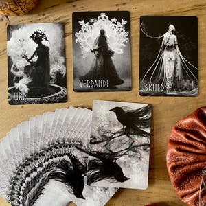 May include: A deck of tarot cards with black and white illustrations of figures and ravens. The cards are fanned out on a wooden surface with a brown leather pouch and a small red stone.