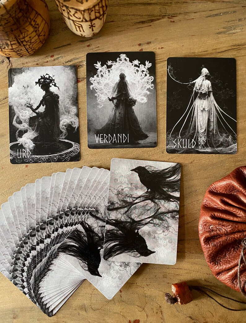 May include: A deck of tarot cards with black and white illustrations of figures and ravens. The cards are fanned out on a wooden surface with a brown leather pouch and a small red stone.