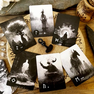 May include: A set of black and white tarot cards with runes and symbols on a wooden table with crystals and a small wooden bowl. The cards are spread out in a circle with a small black hammer in the center.