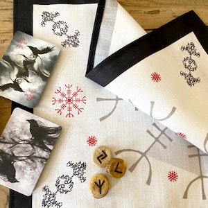 May include: A white cloth with a black border, featuring various Norse symbols and runes in grey and red. There are three wooden runes on the cloth, and two cards with black ravens on a grey background.