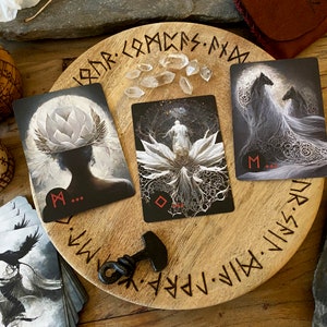 Puede incluir: Tres cartas de tarot colocadas sobre una tabla de madera con runas inscritas alrededor del borde. Las cartas son negras con im&aacute;genes blancas y tienen un s&iacute;mbolo "M" rojo en la esquina inferior. La tabla est&aacute; rodeada de otros objetos, incluyendo un peque&ntilde;o cuenco de madera, un cuchillo y cristales.
