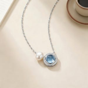 Puede incluir: Un collar de plata con una perla blanca y un colgante redondo con una piedra central azul claro, rodeado de pequeñas piedras transparentes. El collar se muestra sobre una superficie beige claro.