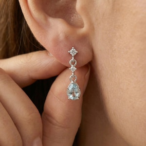 May include: A silver-toned drop earring featuring a small round clear stone at the top, a second smaller round clear stone in the middle, and a larger pear-shaped light blue gemstone at the bottom, all set in a polished metallic frame.