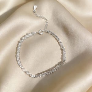 Puede incluir: Una delicada pulsera de plata con un diseño de cadena de doble hebra. La pulsera presenta una pequeña cadena ajustable y un cierre seguro. La joya se exhibe sobre una superficie de satén, destacando sus cualidades reflectantes.