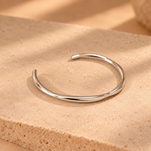 May include: A silver-toned metal cuff bracelet with a smooth, polished finish and a slightly irregular, organic shape. The bracelet features an open design, allowing it to be slipped on and off the wrist with ease, showcasing a reflective surface.