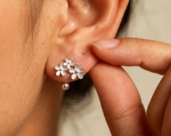 Op de afbeelding: Een zilveren oorsteker met een cluster van drie kleine bloemvormen met getextureerde blaadjes. Een enkele gladde zilveren kraal hangt onderaan het bloemmotief, dat met een standaard metalen sluiting aan de oorlel is bevestigd.
