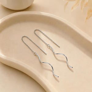 May include: A pair of silver-colored threader earrings. Each earring features a delicate chain that leads to a wavy, elongated design. The earrings are displayed on a light beige, oval-shaped tray.