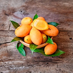 May include: A wooden bowl filled with several oval-shaped, orange-yellow marian plum fruits. The fruits have smooth, thin skins and are interspersed with several pointed, dark green leaves. The bowl is positioned centrally in the frame.