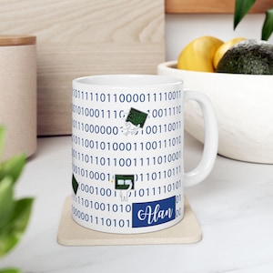 May include: White ceramic mug with a binary code pattern in blue. The mug features a small graphic of a computer chip and the name "Alan" in a script font. The mug sits on a light-colored coaster.