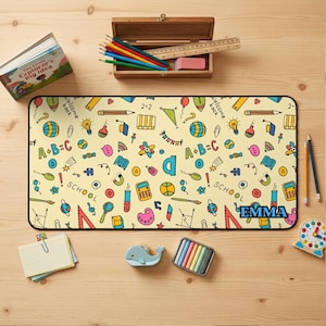 May include: A rectangular desk mat with a cream background featuring a colorful pattern of school-themed icons including calculators, globes, and pencils. The name EMMA is printed in bold blue letters on the bottom right corner of the mat.