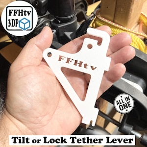May include: A white plastic Tilt or Lock Tether Lever with the text "FFHtv" and "ALL IN ONE". The lever is held in a hand and has a unique shape with a hook and a triangular opening. The product is designed for 3D printing.