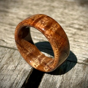 May include: A handcrafted wooden ring with a rich, warm brown color and a glossy finish. The ring showcases the natural wood grain, creating a unique and organic aesthetic. The ring is approximately 0.75 inches wide.