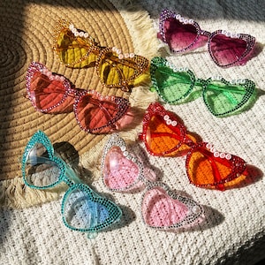 May include: A collection of heart-shaped sunglasses in various colors, including yellow, pink, red, green, and blue. Each pair is embellished with rhinestones and features text across the top of the frames. The sunglasses are arranged on a woven surface.