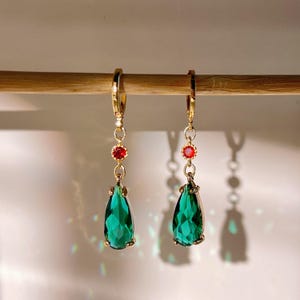 Può includere: Un paio di orecchini pendenti color oro. Ogni orecchino presenta una gemma rossa circolare e una grande gemma verde sfaccettata a forma di goccia. Gli orecchini sono appesi a un tassello di legno su sfondo bianco.