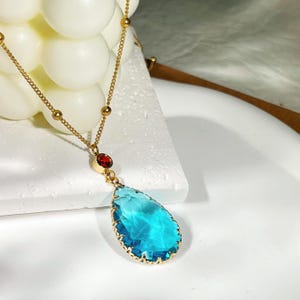 Op de afbeelding: Een goudkleurige ketting met een grote, druppelvormige, lichtblauwe hanger met een goudkleurige rand. De ketting bevat ook een kleine, rode, ovale steen en een fijne ketting.