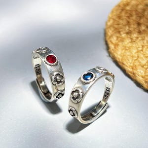 以下が含まれることがあります： 花柄と宝石がアクセントのシルバーリング2つ。1つは赤い宝石、もう1つは青い宝石が付いています。リングは調整可能で、「S925」の刻印入りです。