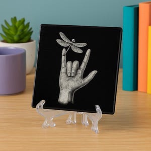 Puede incluir: Azulejo decorativo cuadrado negro con un gesto de la mano y una ilustración de libélula. El azulejo se exhibe en un soporte de plástico transparente. El gesto de la mano es el signo "Te amo".