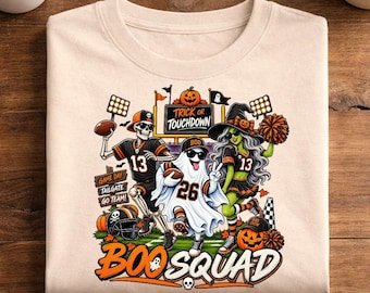 May include: A beige t-shirt featuring a graphic of a skeleton, a ghost, and a green-skinned character dressed in football uniforms. The design includes the text "TRICK OR TOUCHDOWN" and "BOO SQUAD" with footballs, pumpkins, and pom-poms.
