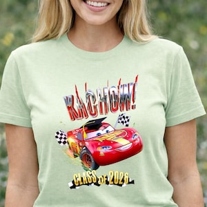 K&ouml;nnte beinhalten: Hellgr&uuml;nes T-Shirt mit einem roten Rennwagen-Motiv, dem Text "KACHOW!" und "CLASS OF 2026". Der Rennwagen hat eine Abschlusskappe und Zielflaggen. Das T-Shirt ist ein kurz&auml;rmeliges Rundhals-Shirt.