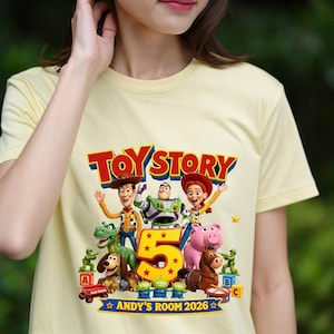 May include: A pale yellow t-shirt featuring a colorful Toy Story 5 graphic. The design includes characters like Woody, Buzz Lightyear, Jessie, Rex, Slinky Dog, Hamm, Bullseye, and green army men, with the text "TOY STORY 5" and "ANDY'S ROOM 2026".