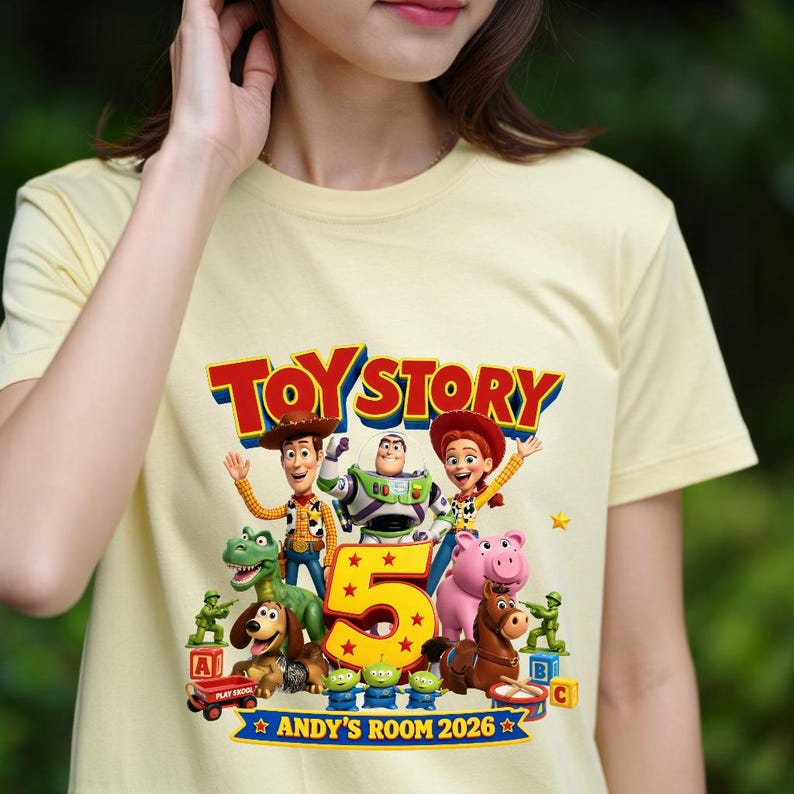 May include: A pale yellow t-shirt featuring a colorful Toy Story 5 graphic. The design includes characters like Woody, Buzz Lightyear, Jessie, Rex, Slinky Dog, Hamm, Bullseye, and green army men, with the text "TOY STORY 5" and "ANDY'S ROOM 2026".