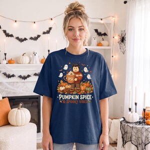 May include: A navy blue t-shirt featuring a graphic of a cartoon character holding a mug labeled PSL, surrounded by pumpkins, autumn leaves, and small ghosts. The text PUMPKIN SPICE & SPOOKY VIBES is printed in white and orange below the illustration.