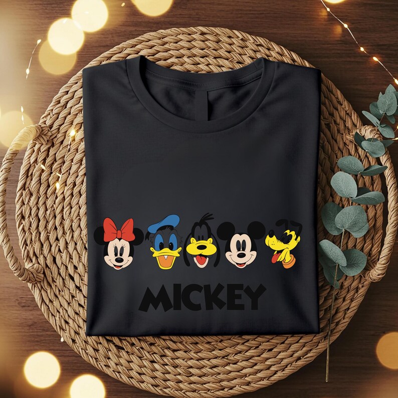 May include: Black t-shirt featuring the faces of Minnie Mouse, Donald Duck, Goofy, Mickey Mouse, and Pluto. The word "MICKEY" is printed in black below the characters. The shirt is folded and placed on a woven basket.