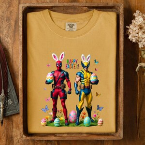 Puede incluir: Camiseta amarilla mostaza con los personajes de Deadpool y Wolverine con orejas de conejo, sosteniendo huevos de Pascua. Las palabras "HAPPY EASTER!" se muestran encima de ellos, con mariposas y huevos de Pascua.