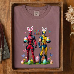 Puede incluir: Sudadera malva con Deadpool y Wolverine disfrazados de conejitos de Pascua, sosteniendo huevos decorados. Las palabras "Happy Easter!" est&aacute;n encima de ellos, con mariposas y flores esparcidas.