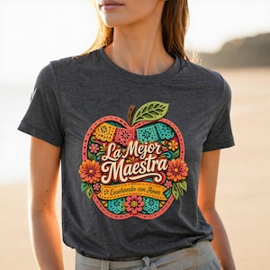 May include: Dark gray t-shirt featuring a colorful apple design with the text "La Mejor Maestra" and "Enseñando con Amor". The apple design includes floral and decorative elements in various colors, including pink, orange, and turquoise.