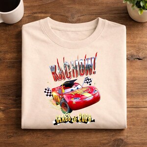 K&ouml;nnte beinhalten: Beiges Sweatshirt mit einer Grafik von Lightning McQueen, einem roten Rennwagen aus dem Film Cars, der eine Abschlusskappe tr&auml;gt. Der Text "KACHOW!" und "Class of 2026" sind ebenfalls abgebildet.