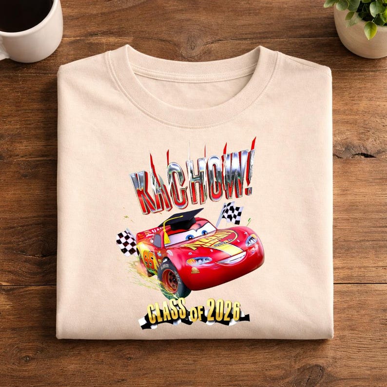 K&ouml;nnte beinhalten: Beiges Sweatshirt mit einer Grafik von Lightning McQueen, einem roten Rennwagen aus dem Film Cars, der eine Abschlusskappe tr&auml;gt. Der Text "KACHOW!" und "Class of 2026" sind ebenfalls abgebildet.