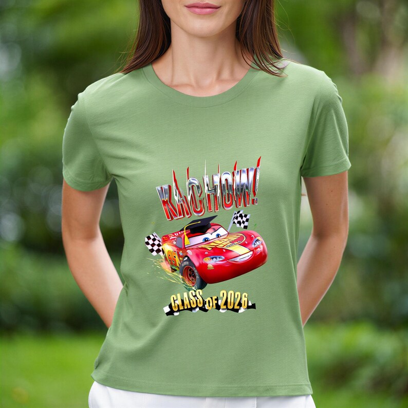 K&ouml;nnte beinhalten: Gr&uuml;nes T-Shirt mit einem roten Cartoon-Rennwagen, dem Text "KACHOW!" und "CLASS OF 2026". Das Auto tr&auml;gt eine Abschlusskappe und hat karierte Flaggen. Das T-Shirt hat eine einfarbige gr&uuml;ne Farbe.