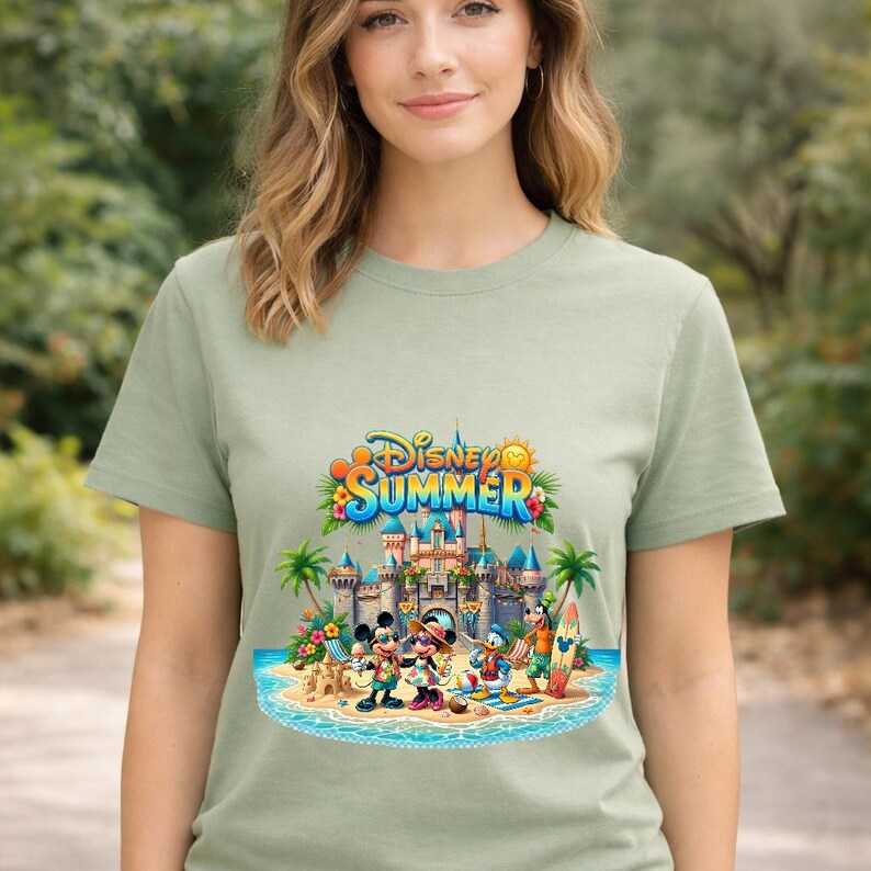 May include: A sage green t-shirt featuring a colorful graphic of Disney characters including Mickey Mouse, Minnie Mouse, Donald Duck, and Goofy at a beach with a castle. The text "Disney SUMMER" is printed in blue and yellow above the castle scene.