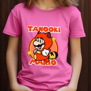 May include: A bright pink short-sleeved t-shirt featuring a graphic of Tanooki Mario in an orange suit with a striped tail. The design includes yellow stars, an orange circular background, and the text TANOOKI MARIO in bold white letters.