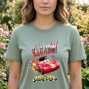 K&ouml;nnte beinhalten: Salbeigr&uuml;nes T-Shirt mit einer roten Rennwagen-Grafik und dem Text "KACHOW!" in einer chromartigen Schrift. Die Grafik enth&auml;lt den Text "CLASS OF 2026" und karierte Flaggen. Das T-Shirt hat einen Rundhalsausschnitt.