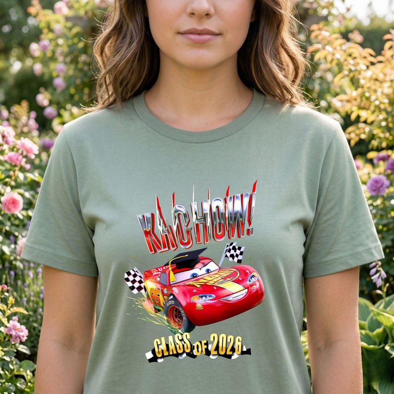 K&ouml;nnte beinhalten: Salbeigr&uuml;nes T-Shirt mit einer roten Rennwagen-Grafik und dem Text "KACHOW!" in einer chromartigen Schrift. Die Grafik enth&auml;lt den Text "CLASS OF 2026" und karierte Flaggen. Das T-Shirt hat einen Rundhalsausschnitt.