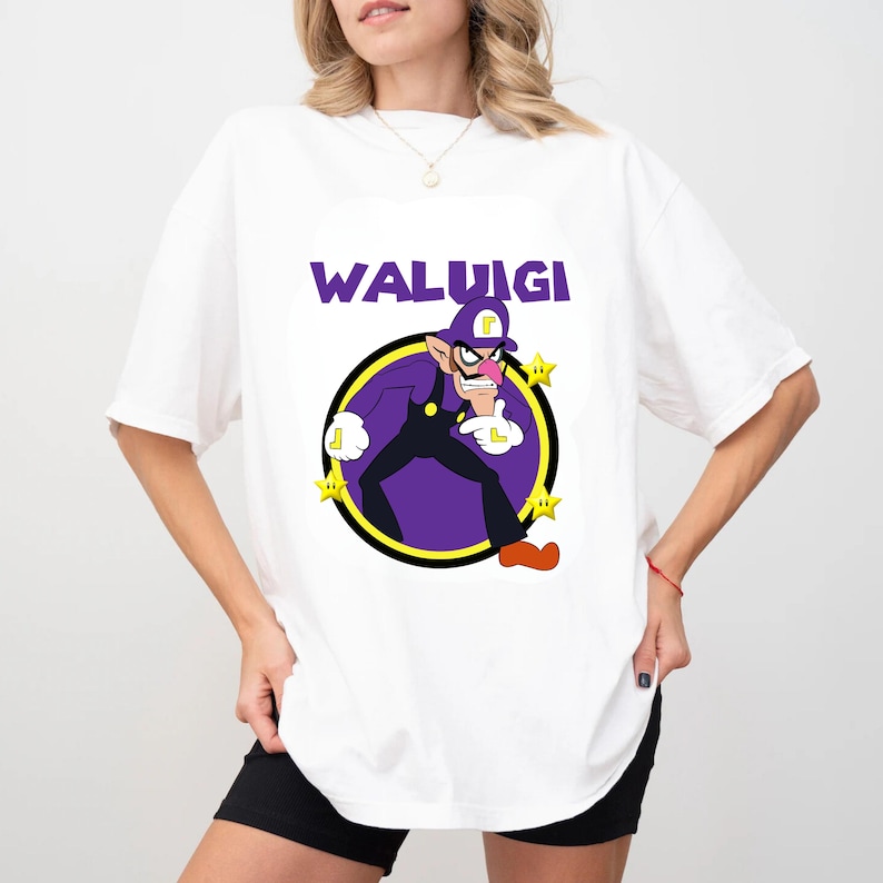 May include: A white short-sleeved t-shirt featuring a graphic of the character Waluigi in a purple outfit with a yellow L on his hat and gloves. The text WALUIGI is printed in purple above the character, surrounded by three small yellow stars.