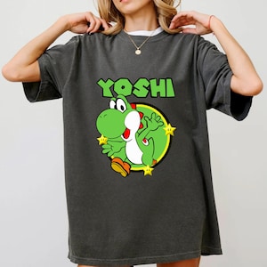 May include: A charcoal gray short-sleeve t-shirt featuring a graphic print of the green character Yoshi from Super Mario. The design includes the text YOSHI in green letters above the character, who is surrounded by three yellow stars.