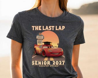May include: A dark heather gray t-shirt featuring a graphic of a red race car wearing a graduation cap. The text reads THE LAST LAP, SENIOR 2027, RADIATOR SPRINGS, and ROUTE 66. The design includes a sunset background and a road leading to a sign.