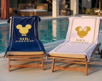 May include: Two personalized beach chair covers. One is navy blue with a gold Mickey Mouse design and the text "MR. SMITH". The other is light pink with a Minnie Mouse design and the text "MRS. SMITH".