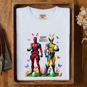 Puede incluir: Camiseta blanca con Deadpool y Wolverine disfrazados de conejos de Pascua, sosteniendo huevos decorados. Las palabras "HAPPY EASTER!" est&aacute;n impresas encima de ellos, con mariposas y huevos de Pascua. La camiseta tiene el texto "RIGHTEOUS PRINT" en la parte superior izquierda.