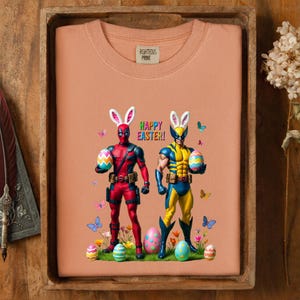 Puede incluir: Sudadera color melocot&oacute;n con Deadpool y Wolverine con orejas de conejito de Pascua, sosteniendo huevos decorados. Las palabras "Happy Easter!" est&aacute;n encima de los personajes. Mariposas y huevos de Pascua est&aacute;n esparcidos alrededor de los personajes. La etiqueta dice "Righteous Print".