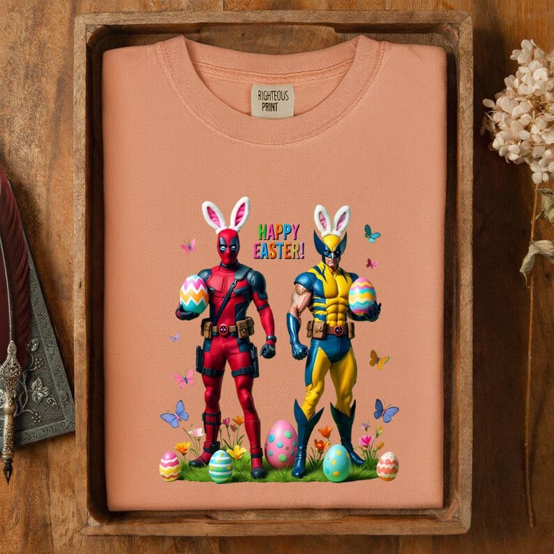Puede incluir: Sudadera color melocot&oacute;n con Deadpool y Wolverine con orejas de conejito de Pascua, sosteniendo huevos decorados. Las palabras "Happy Easter!" est&aacute;n encima de los personajes. Mariposas y huevos de Pascua est&aacute;n esparcidos alrededor de los personajes. La etiqueta dice "Righteous Print".