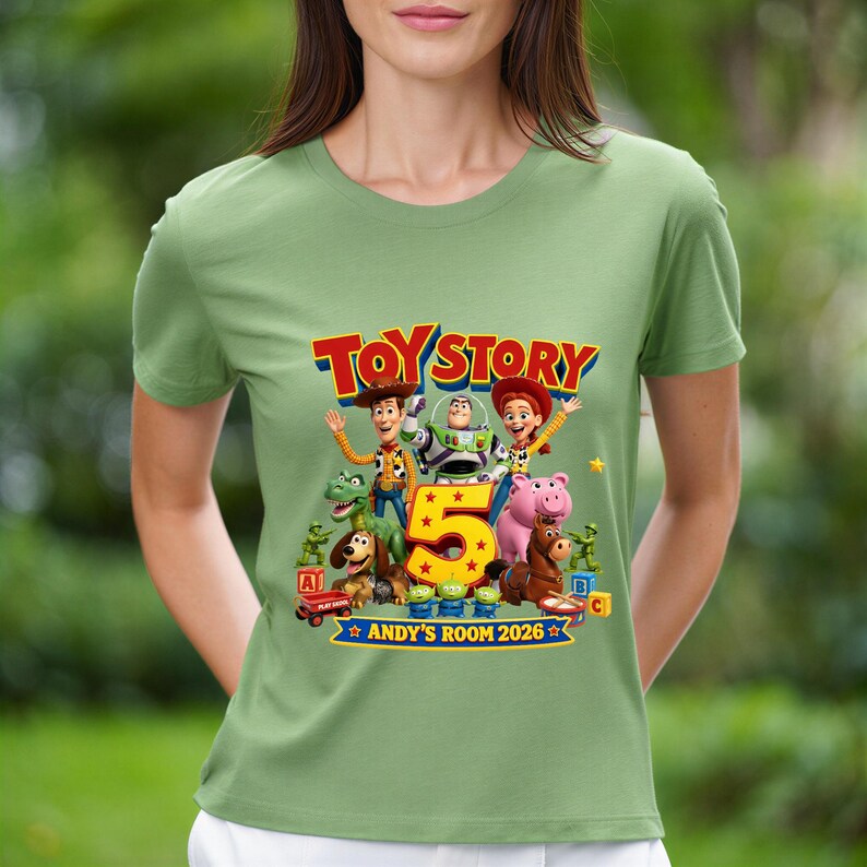 May include: A sage green t-shirt featuring a colorful Toy Story graphic with Woody, Buzz Lightyear, Jessie, and other characters surrounding a large yellow number 5. The text reads Toy Story, 5, and Andy's Room 2026 with small stars.