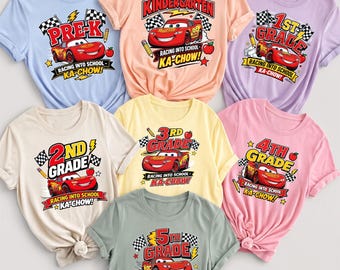 May include: A collection of seven short-sleeved t-shirts in various pastel colors, each featuring a graphic of a red race car, checkered flags, a pencil, an apple, and text indicating school grade levels from Pre-K to 5th Grade with the phrase KA-CHOW!