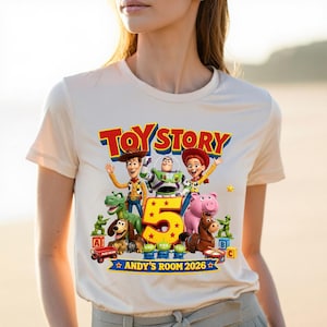 May include: A cream-colored t-shirt featuring a colorful Toy Story graphic with characters Woody, Buzz Lightyear, Jessie, Rex, Slinky Dog, Hamm, Bullseye, and green army men. The design includes a large yellow number 5 and text reading Toy Story Andy's Room 2026.