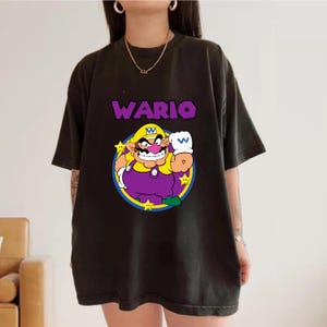 May include: A black short-sleeve t-shirt featuring a graphic of the character Wario in a yellow hat, purple overalls, and white gloves. The character is centered within a blue and yellow circular frame with yellow stars. The text WARIO is printed in purple above the graphic.